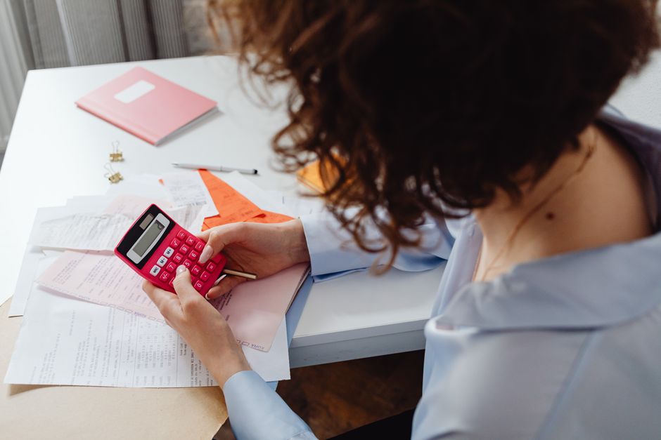 A person sits at a clean, bright desk using a calculator next to a small model of a house, illustrating the financial calculations involved in real estate commissions.