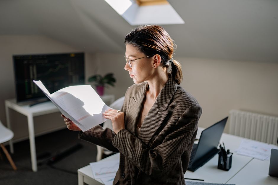 A person stands thoughtfully in a bright, empty room holding a folder of documents, contemplating the space and conveying the serious, technical knowledge required for real estate transactions.